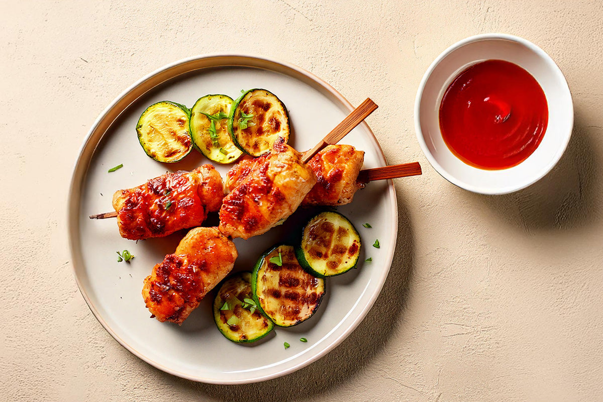 Gegrillte Hähnchenspieße mit Zucchinischeiben auf weißem Teller, daneben Schale mit roter Sauce