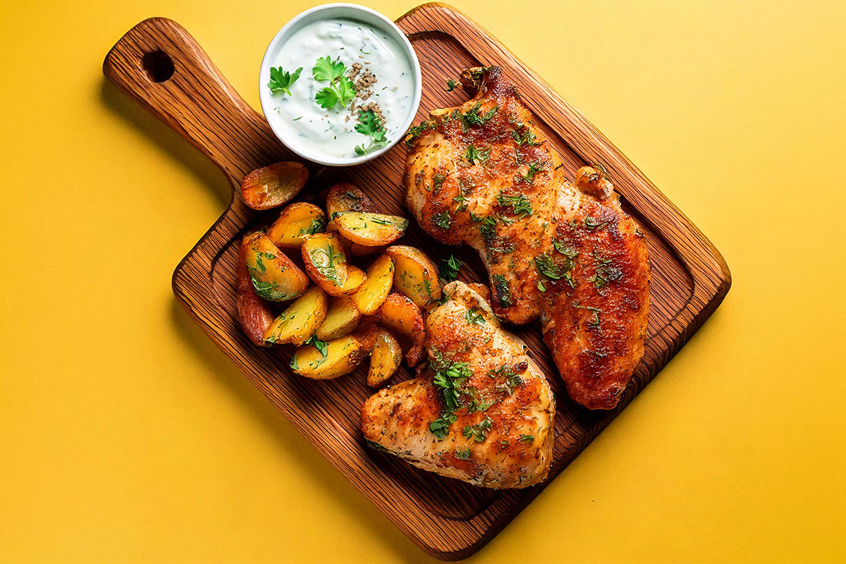 Gegrillte Hähnchensteaks mit frischen Kräutern, Kartoffelecken und einem Dip auf einem Holzbrett vor gelbem Hintergrund