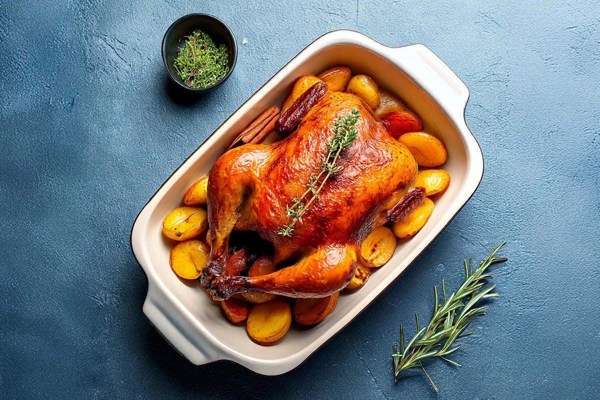 Gegrillter Entenbraten mit knuspriger Haut auf einem Bett aus gebackenen Kartoffeln in einer weißen Auflaufform, garniert mit frischem Thymian und Zimtstangen, daneben Rosmarinzweig und kleine Schale mit Kräutern