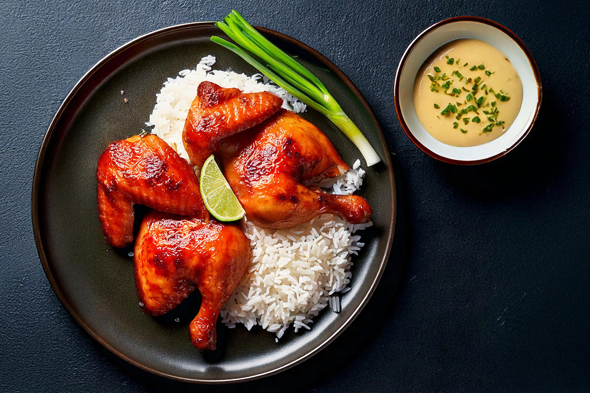 Gegrilltes asiatisches Stubenküken mit weißem Reis, Limettenscheibe und Frühlingszwiebeln auf dunklem Teller, daneben Schale mit Sauce und Kräutern