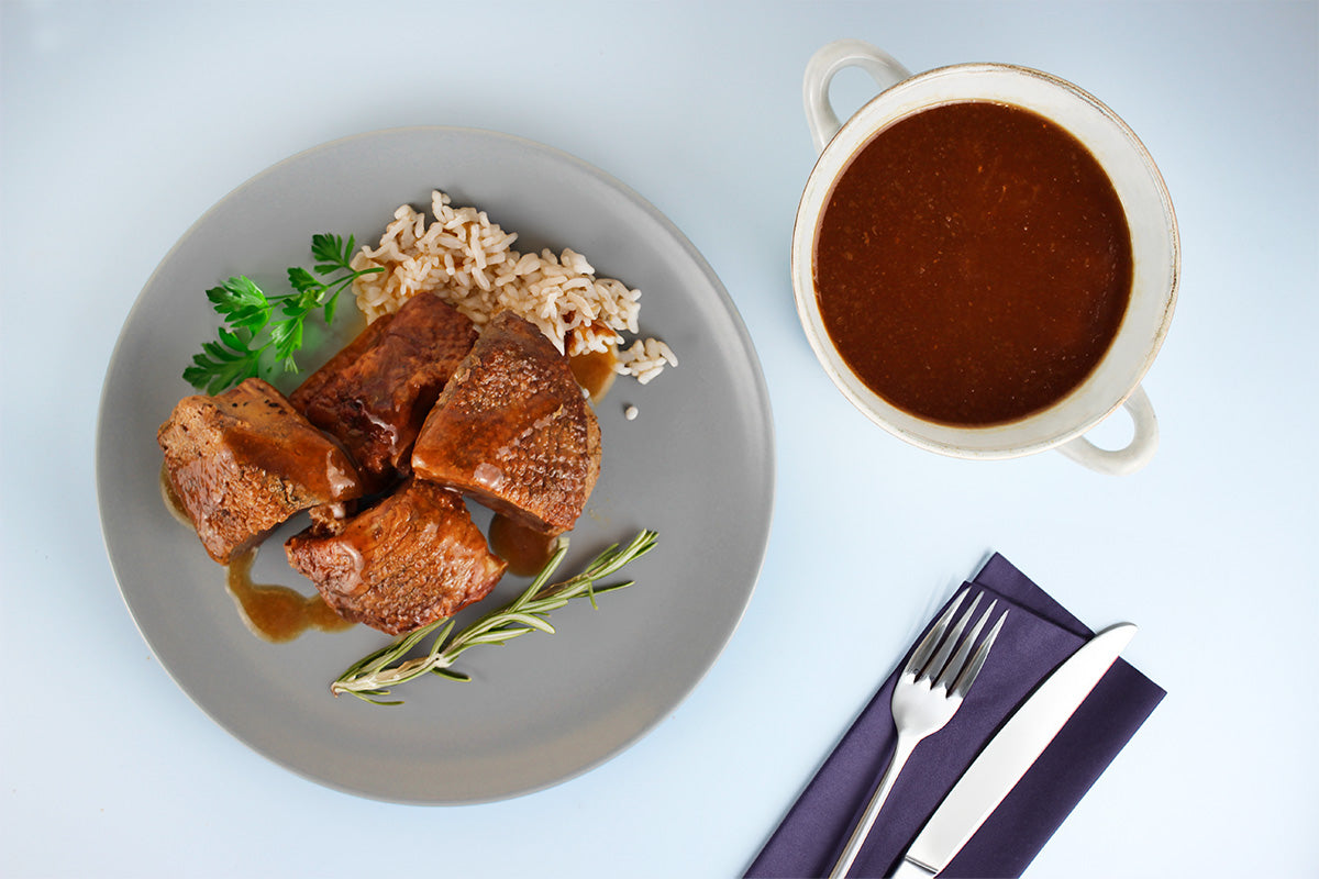 Vier Stücke gebratene Gans mit brauner Sauce, Reis, Rosmarinzweig und Petersilie auf grauem Teller, daneben Schale mit brauner Sauce und Besteck auf dunkelblauem Serviette