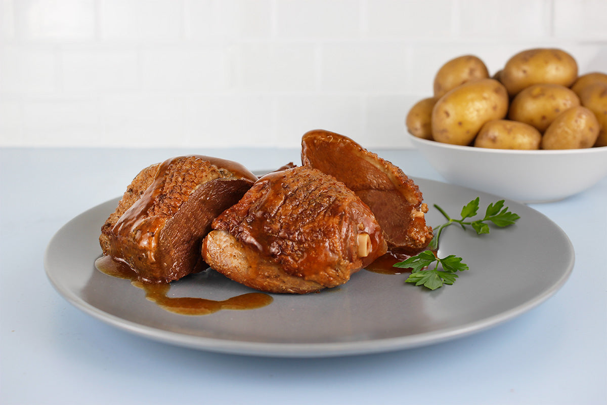 Drei Stücke gebratene Gans mit brauner Sauce auf grauem Teller, daneben Petersilienzweig und Schüssel mit kleinen Kartoffeln