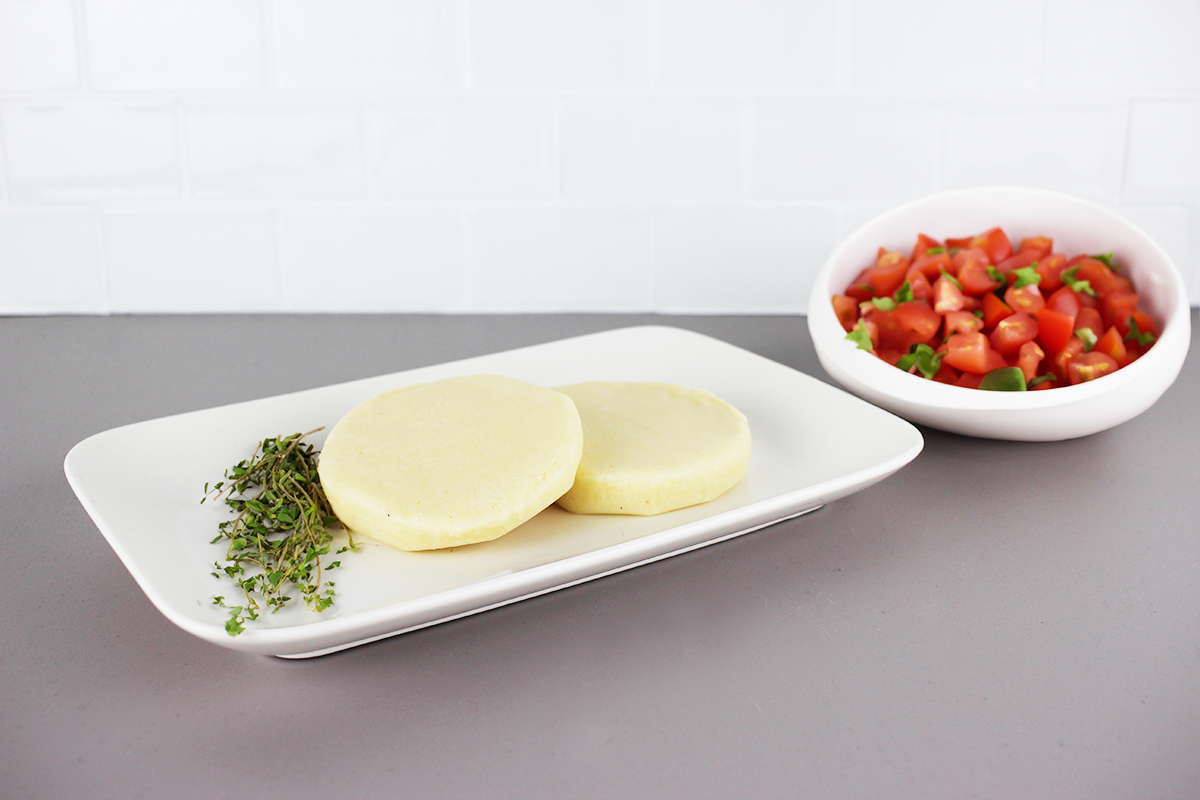 Zwei Scheiben naturbelassener Grillkäse auf weißem Teller mit frischem Thymian, daneben Schale mit Tomatenwürfeln und Basilikum