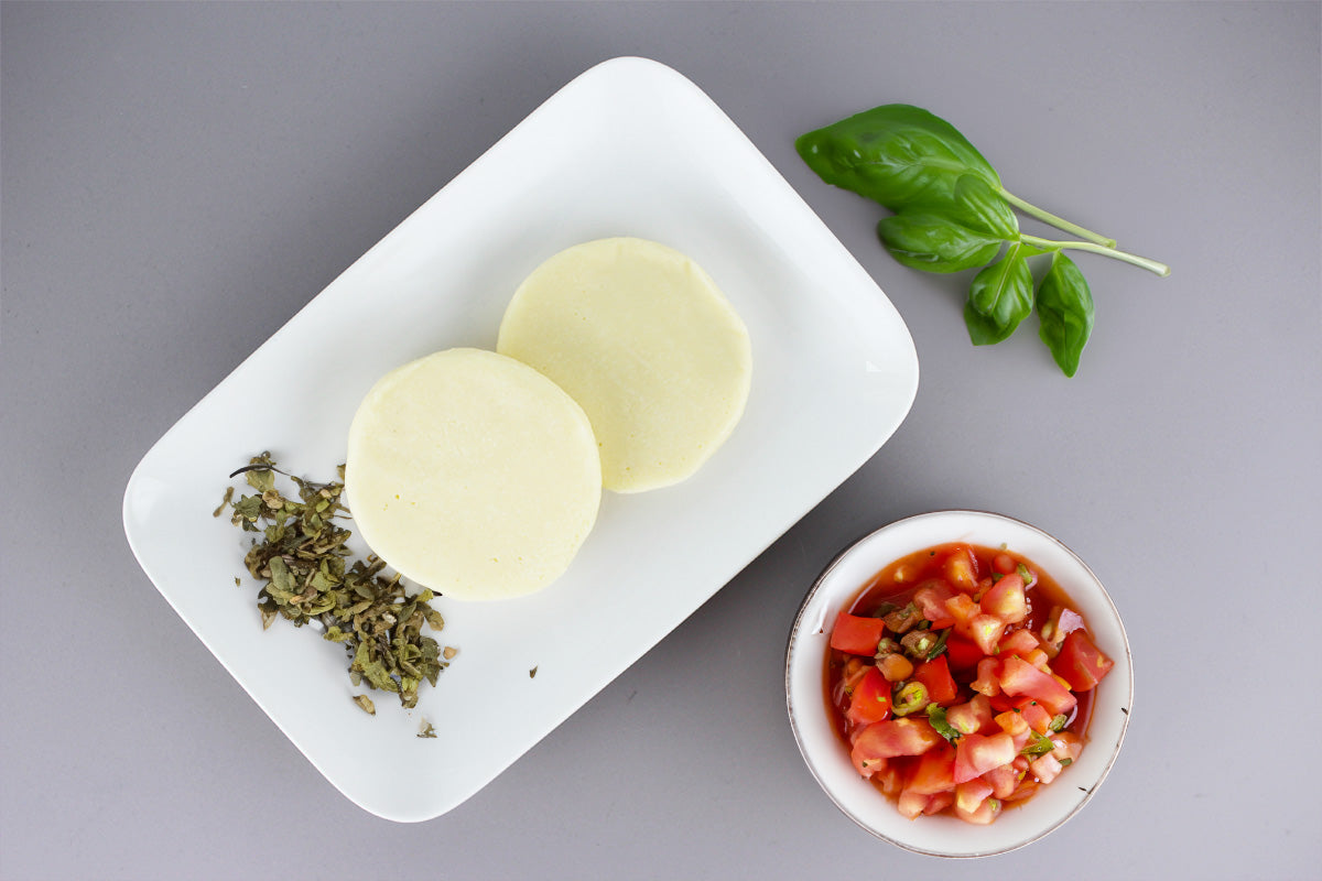 Zwei runde Scheiben Grillkäse auf weißem Teller mit getrockneten Kräutern, daneben Schale mit Tomatenwürfeln und Basilikumblätter