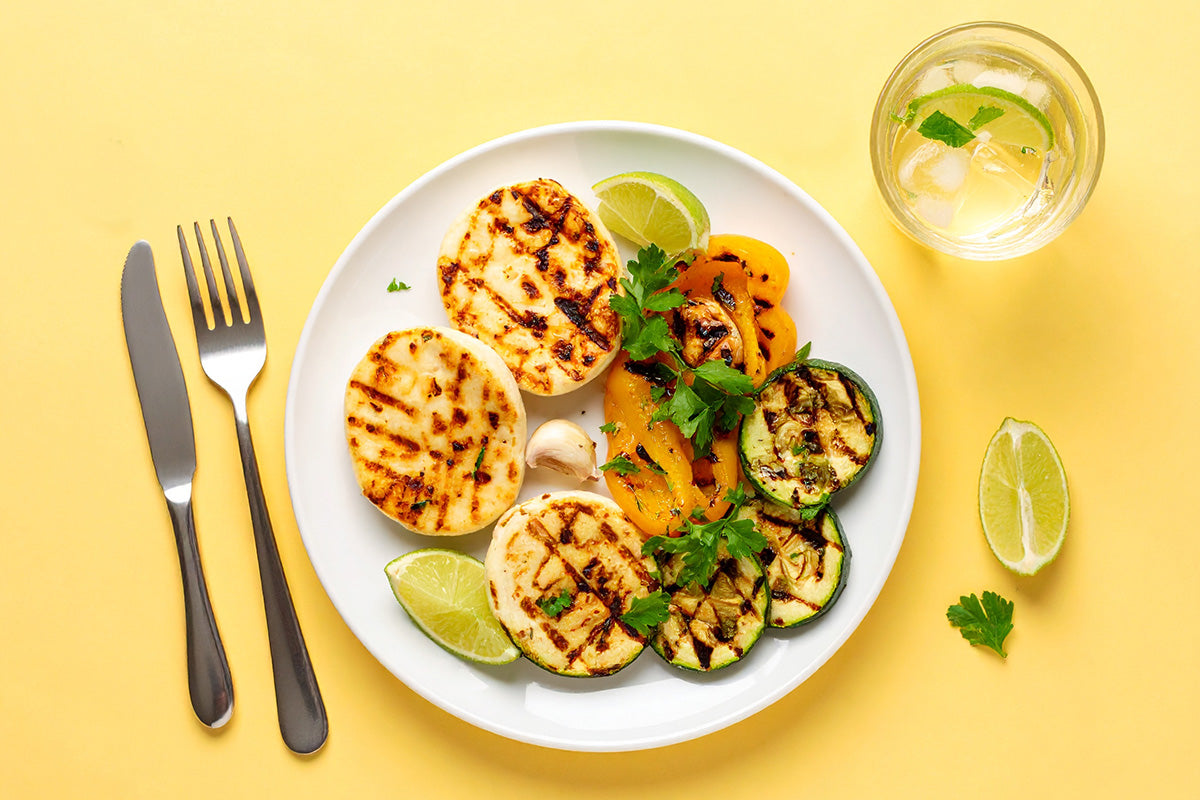 Gegrillter Käse auf weißem Teller mit gegrilltem Gemüse wie Zucchini und Paprika, Limettenspalten und frischer Petersilie, daneben Besteck und ein Glas mit Getränk