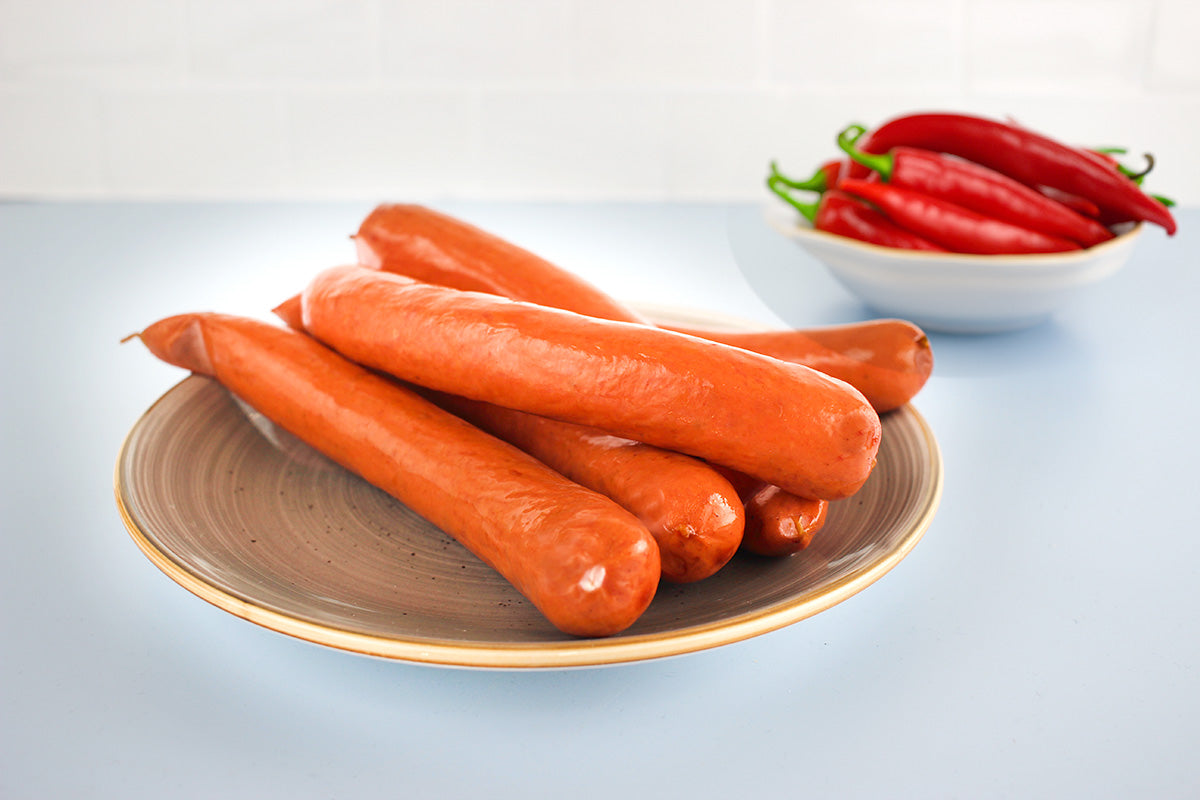 Ein Teller mit fünf orangefarbenen Würstchenauf hellblauen Untergrund, im Hintergrund eine Schale mit roten Chili Schoten