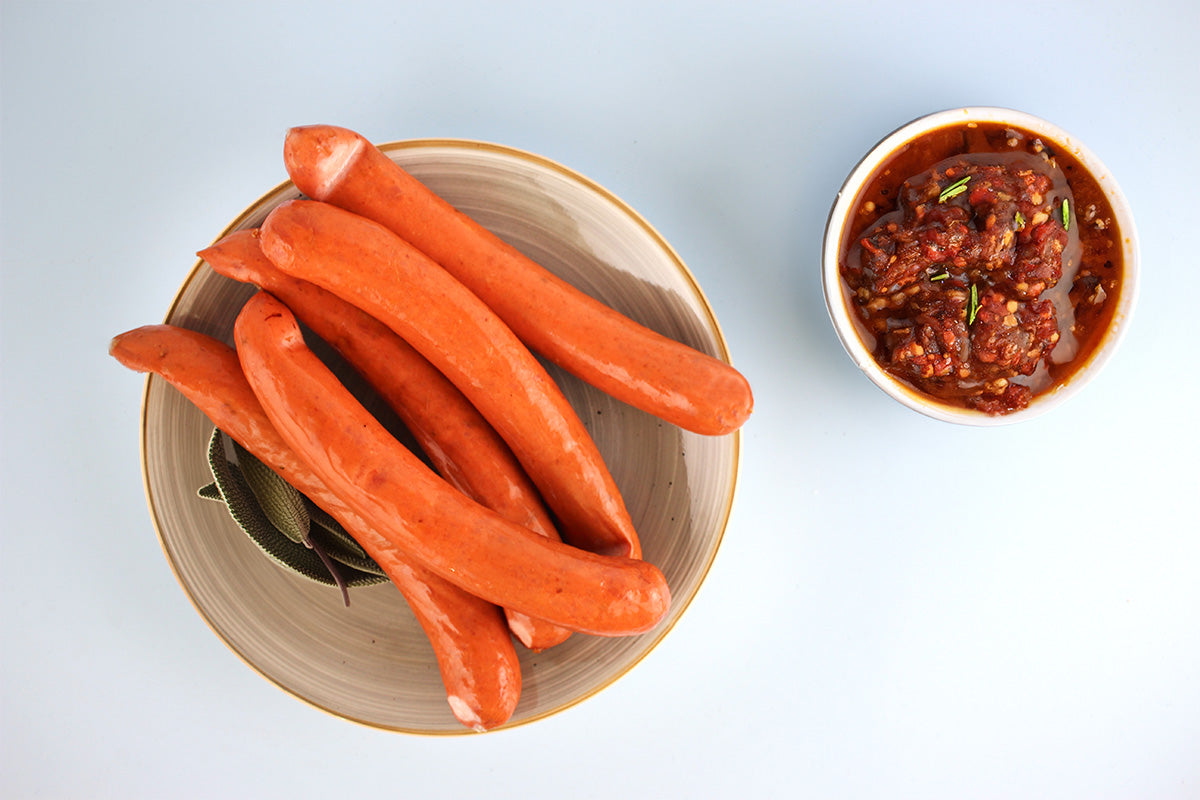 Ein Teller mit fünf orangefarbenen Würstchen neben einer Schüssel mit pikanter roter Sauce mit Kräutern, vor einem hellen Hintergrund.