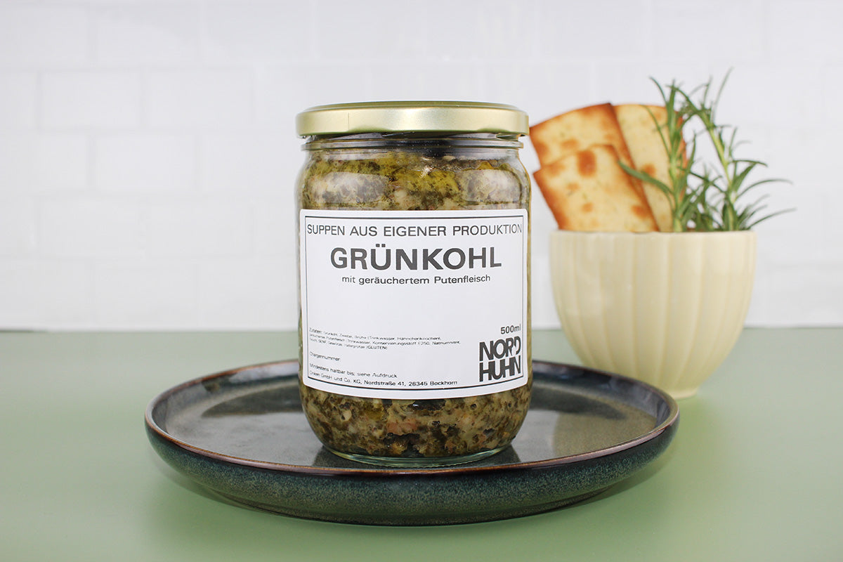 Ein Glas Grünkohl mit geräuchertem Putenfleisch von Nordhuhn auf einem dunklen Teller, daneben eine Tasse mit Brot und Rosmarin