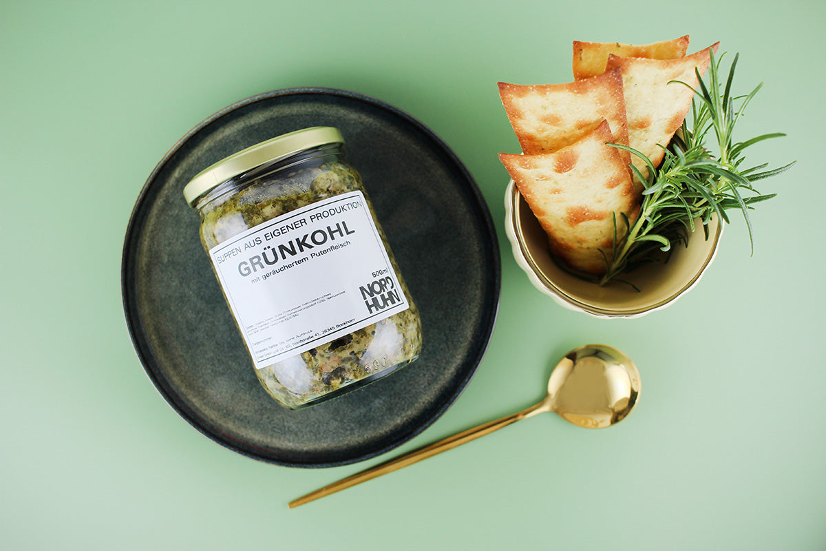 Glas mit Grünkohl auf dunklem Teller, daneben goldener Löffel und Schale mit knusprigem Brot und Rosmarinzweig