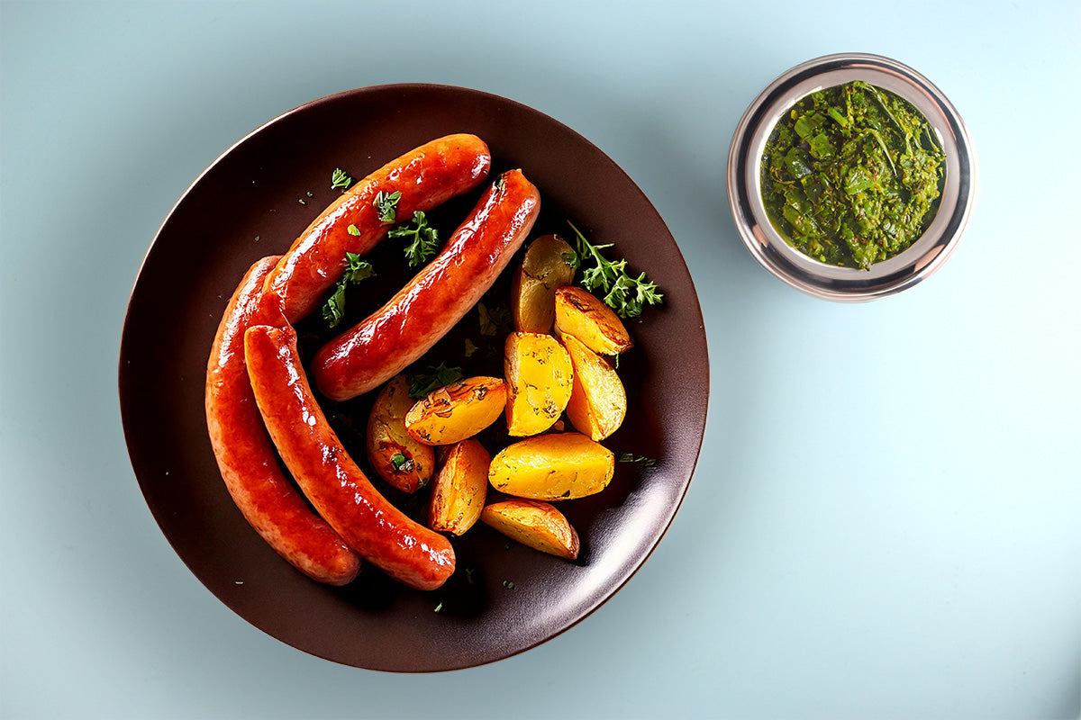 Vier gebratene Würste mit gebratenen Kartoffelspalten auf braunem Teller, daneben Schüssel mit Grünkohl