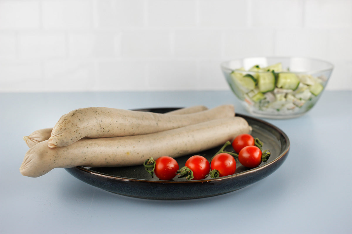 Drei helle Geflügel-Gyrosbratwürste auf schwarzem Teller mit Kirschtomaten, im Hintergrund Schüssel mit Gurkensalat