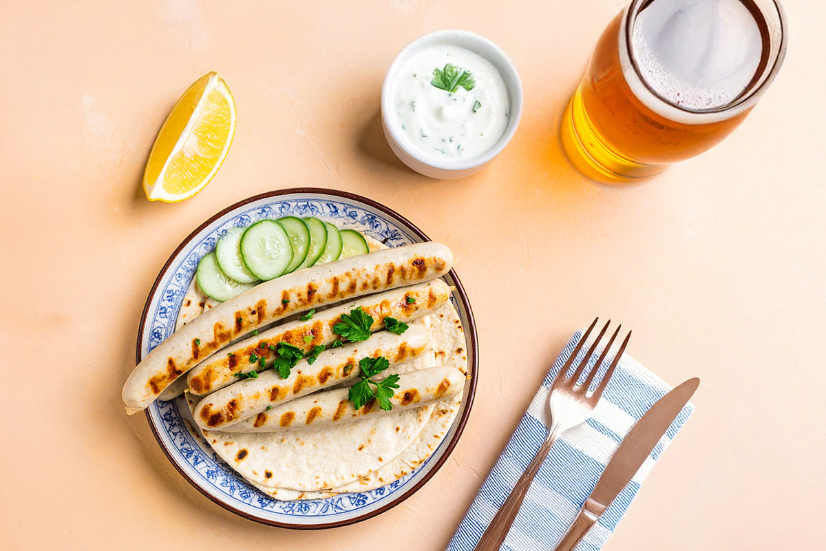 Gegrillte Gyrosbratwürste auf Fladenbrot mit Gurkenscheiben und Petersilie, dazu Joghurt-Dip, Zitronenschnitz und Glas Bier