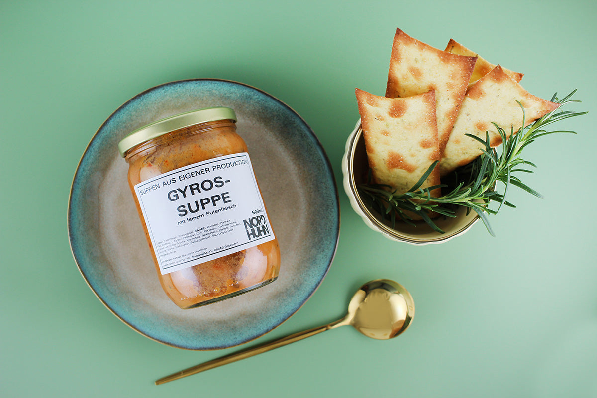 Ein Glas Gyrossuppe mit feinem Putenfleisch auf einem blauen Teller, daneben goldener Löffel und Schale mit knusprigen Fladenbrotstücken und Rosmarin auf grünem Hintergrund