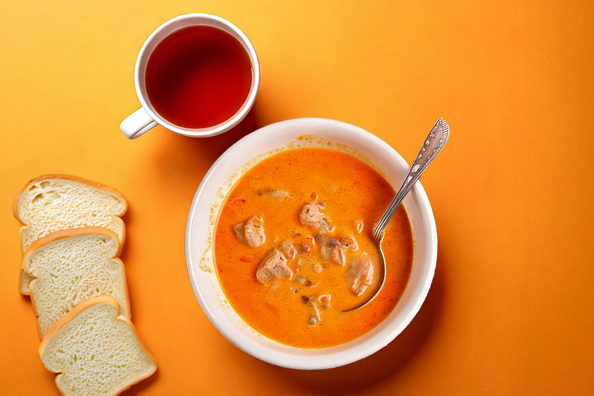 Weiße Schüssel mit Gyrossuppe, Löffel darin, daneben Tasse mit roter Flüssigkeit und drei Scheiben Weißbrot auf orangefarbenem Hintergrund