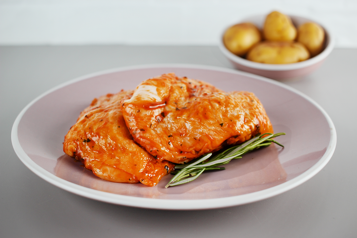 Zwei mit Barbecue-Sauce marinierte Hähnchensteaks auf einem rosa Teller mit einem Rosmarinzweig, im Hintergrund eine Schale mit kleinen Kartoffeln