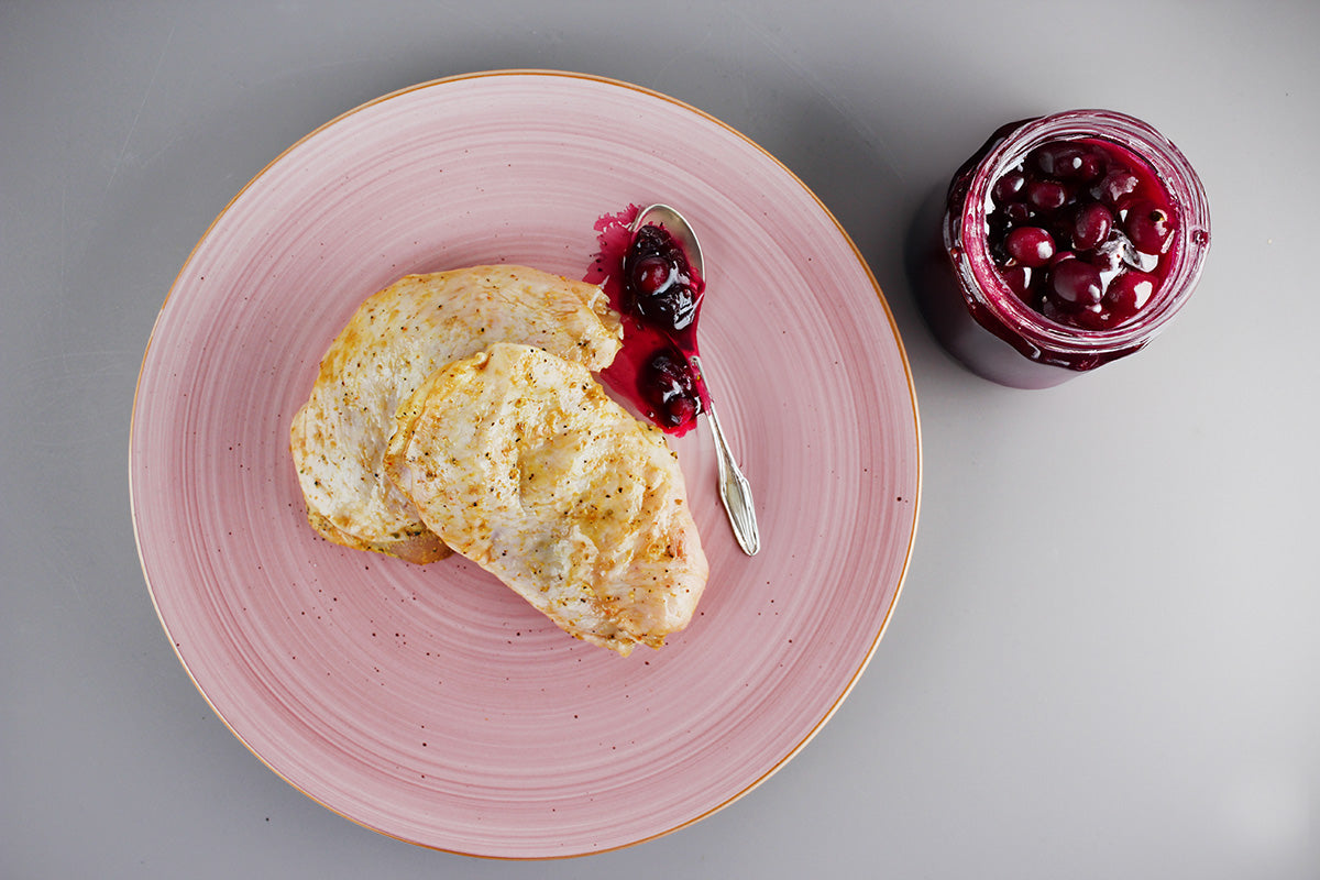 Zwei gebratene Hähnchensteaks auf einem rosa Teller mit einem Klecks Kirschsoße und einem Löffel daneben, daneben ein Glas mit Kirschsoße