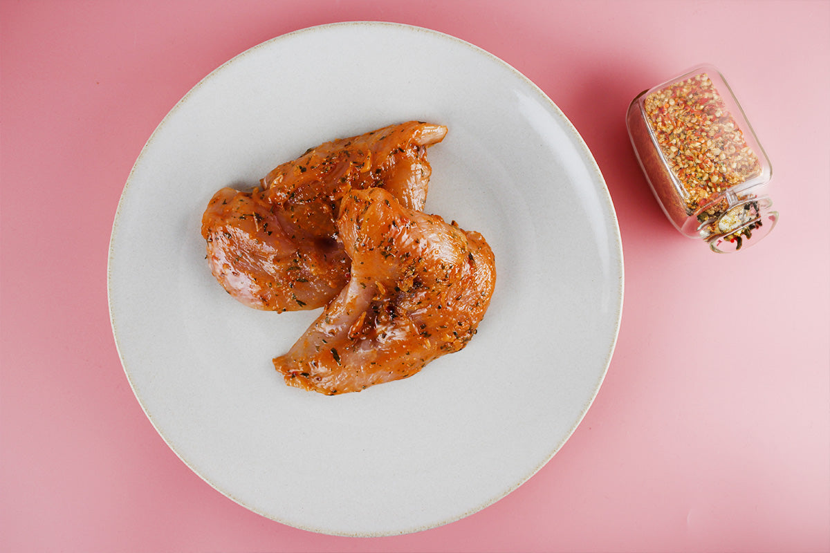 Zwei marinierte Hähnchenbrustfilets mit BBQ-Gewürzen auf weißem Teller, daneben Gewürzglas auf rosa Untergrund