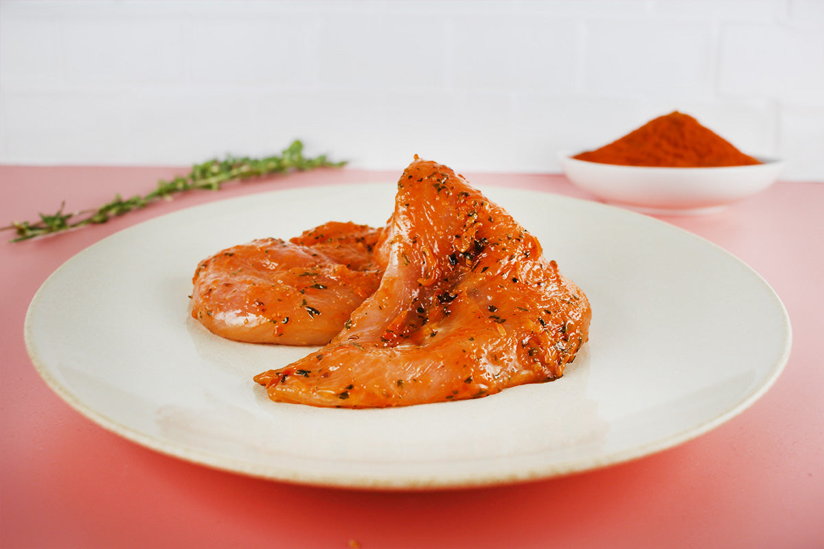 Zwei marinierte Hähnchenbrustfilets mit BBQ-Gewürzen auf weißem Teller, im Hintergrund Thymianzweig und Schale mit Paprikapulver