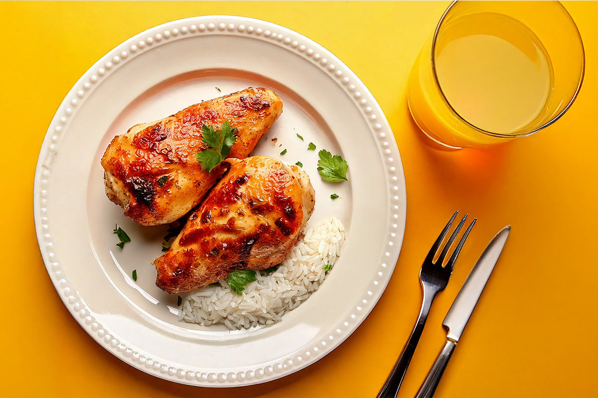 Gegrillte BBQ-Hähnchenbrustfilets auf weißem Teller mit Reis und Petersilie, daneben Glas Orangensaft, Gabel und Messer auf gelbem Untergrund