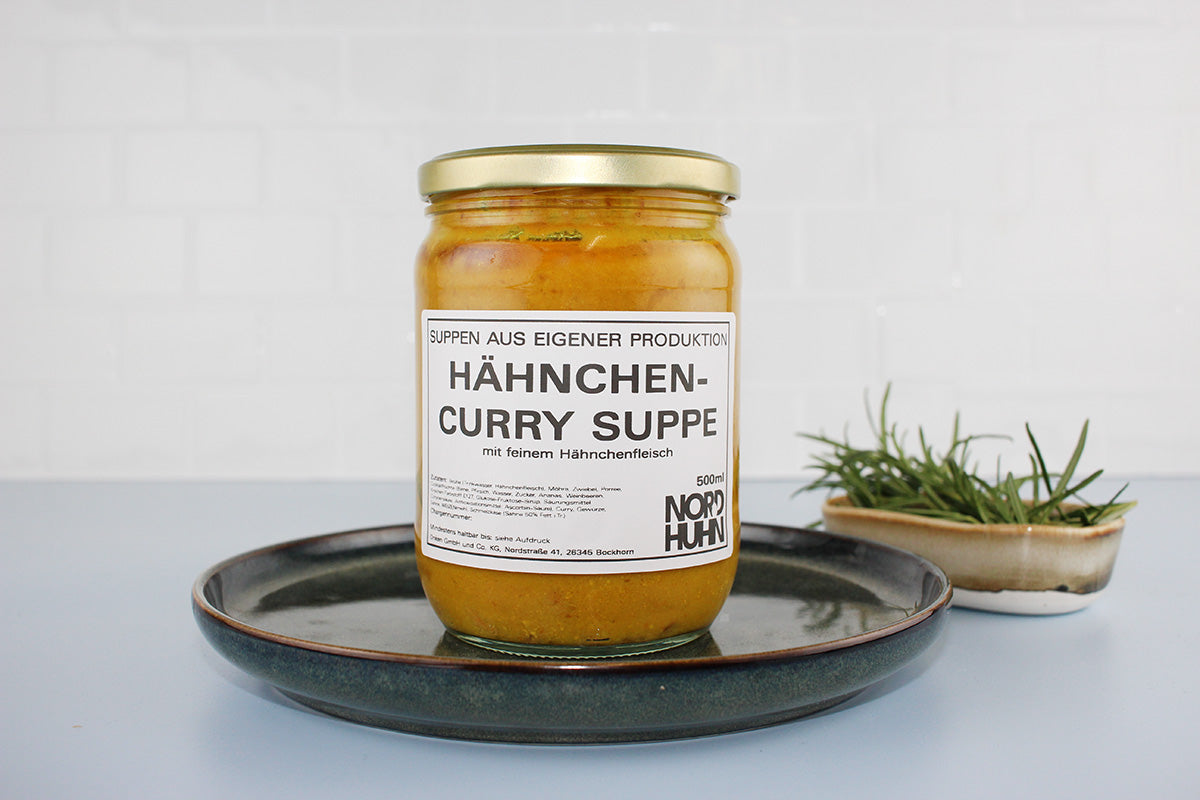 Glas mit Hähnchen-Curry-Suppe von Nordhuhn, 500 ml, auf dunklem Teller, daneben Schale mit frischem Rosmarin
