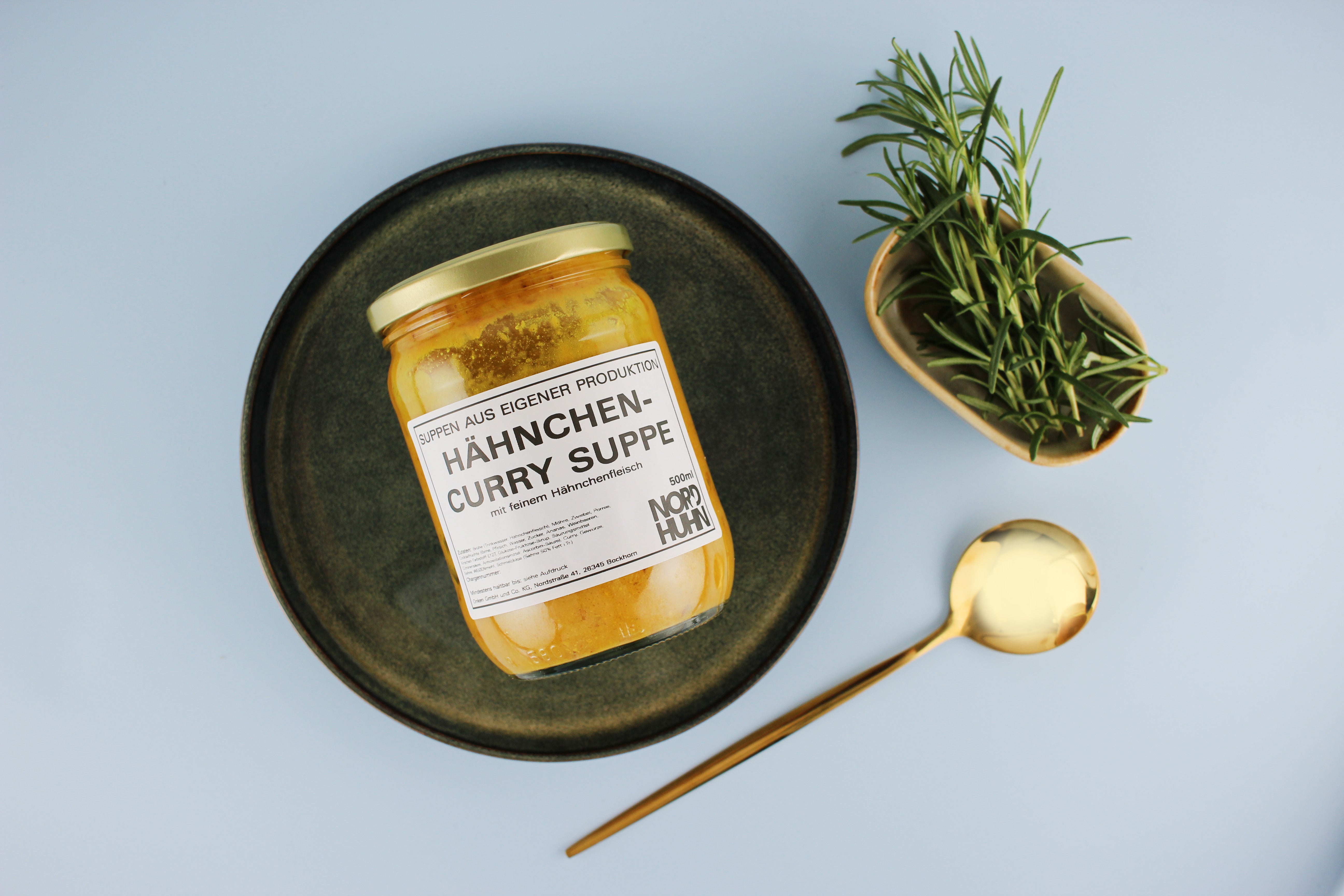 Glas mit Hähnchen-Curry-Suppe von Nordhuhn, 500 ml, auf dunklem Teller, daneben Schale mit frischem Rosmarin