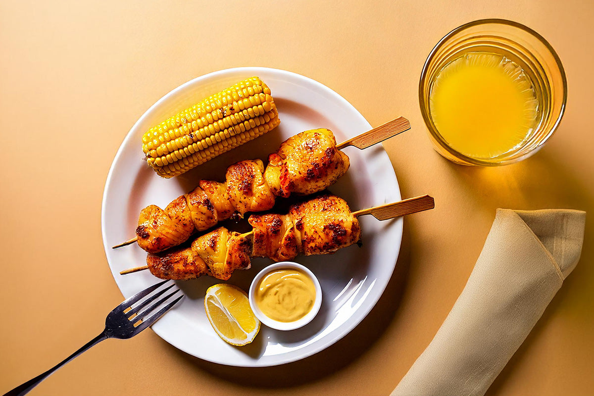 Zwei Curry-Hähnchenfiletspieße auf weißem Teller mit Maiskolben, Zitronenschnitz und Senfsauce, daneben Glas Orangensaft und beige Serviette