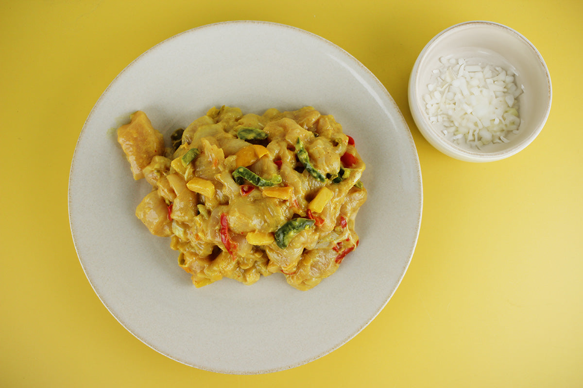 Hähnchenfiletpfanne mit buntem Gemüse und cremiger Sauce auf weißem Teller, daneben kleine Schale mit gehackten Zwiebeln auf gelbem Untergrund