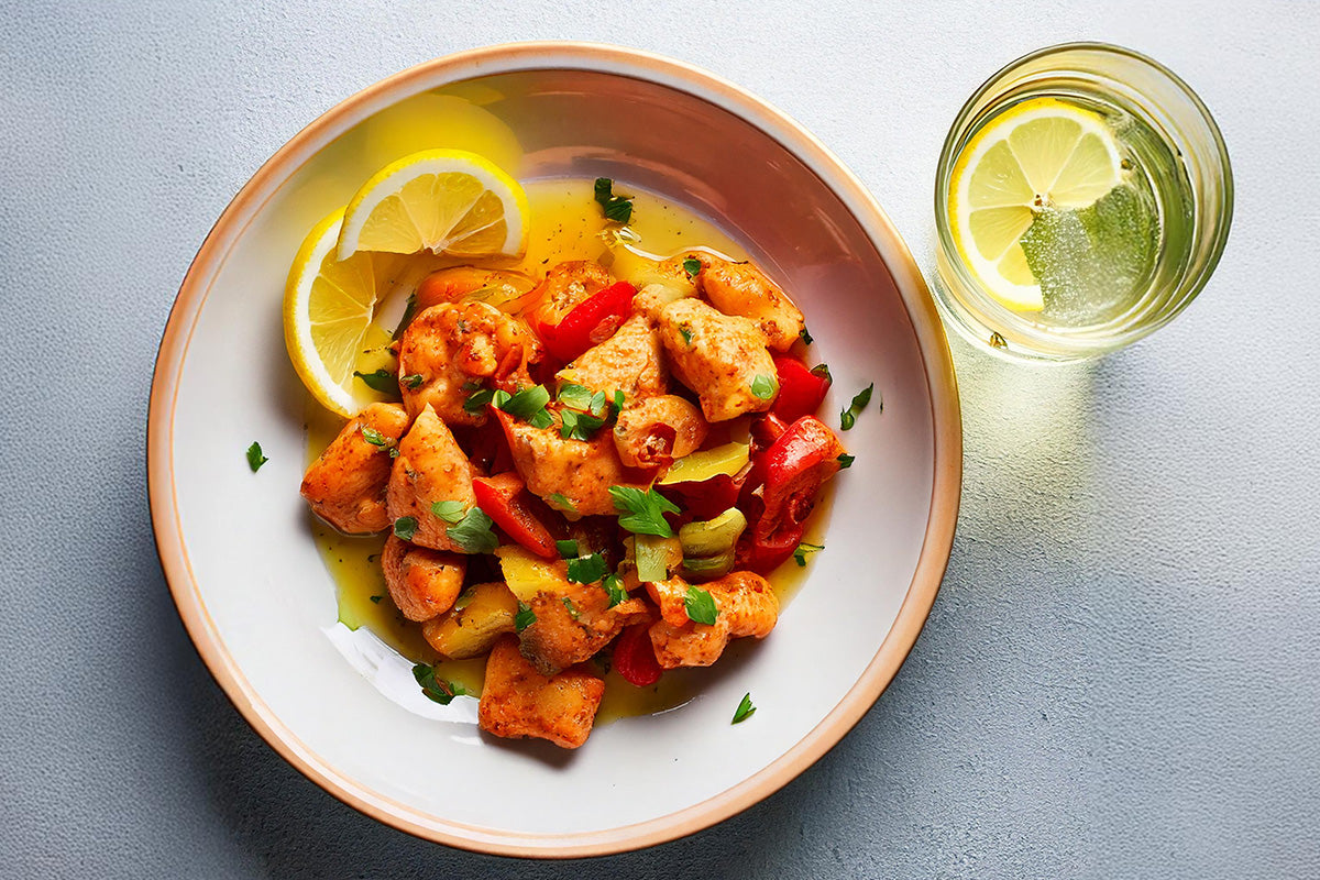 Hähnchenfiletpfanne mit roten und gelben Paprikastücken, Ananasstücken, frischer Petersilie und Zitronenscheiben auf weißem Teller, daneben Glas Wasser mit Zitronenscheibe