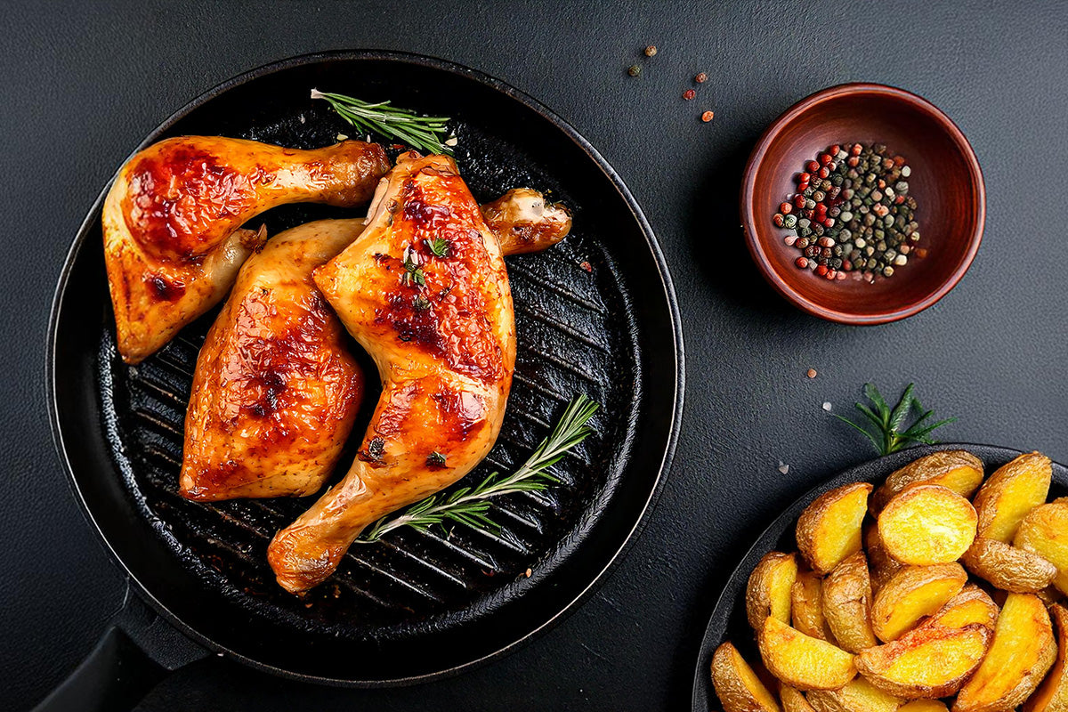 Drei gebratene Hähnchenschenkel mit Rosmarin in einer Grillpfanne, daneben eine Schale mit buntem Pfeffer und eine Pfanne mit gebackenen Kartoffelspalten