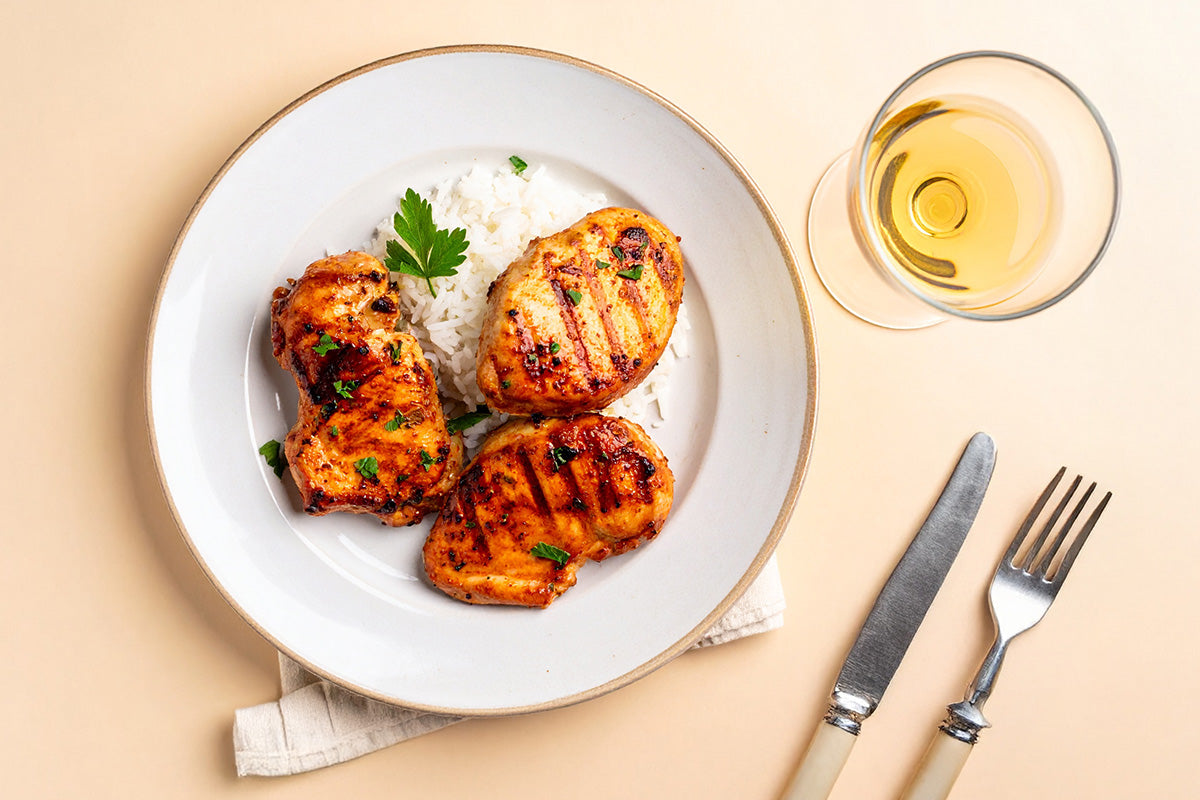 Drei gegrillte Hähnchensteaks mit Reis und Petersilie auf weißem Teller, daneben Messer, Gabel und Glas Weißwein
