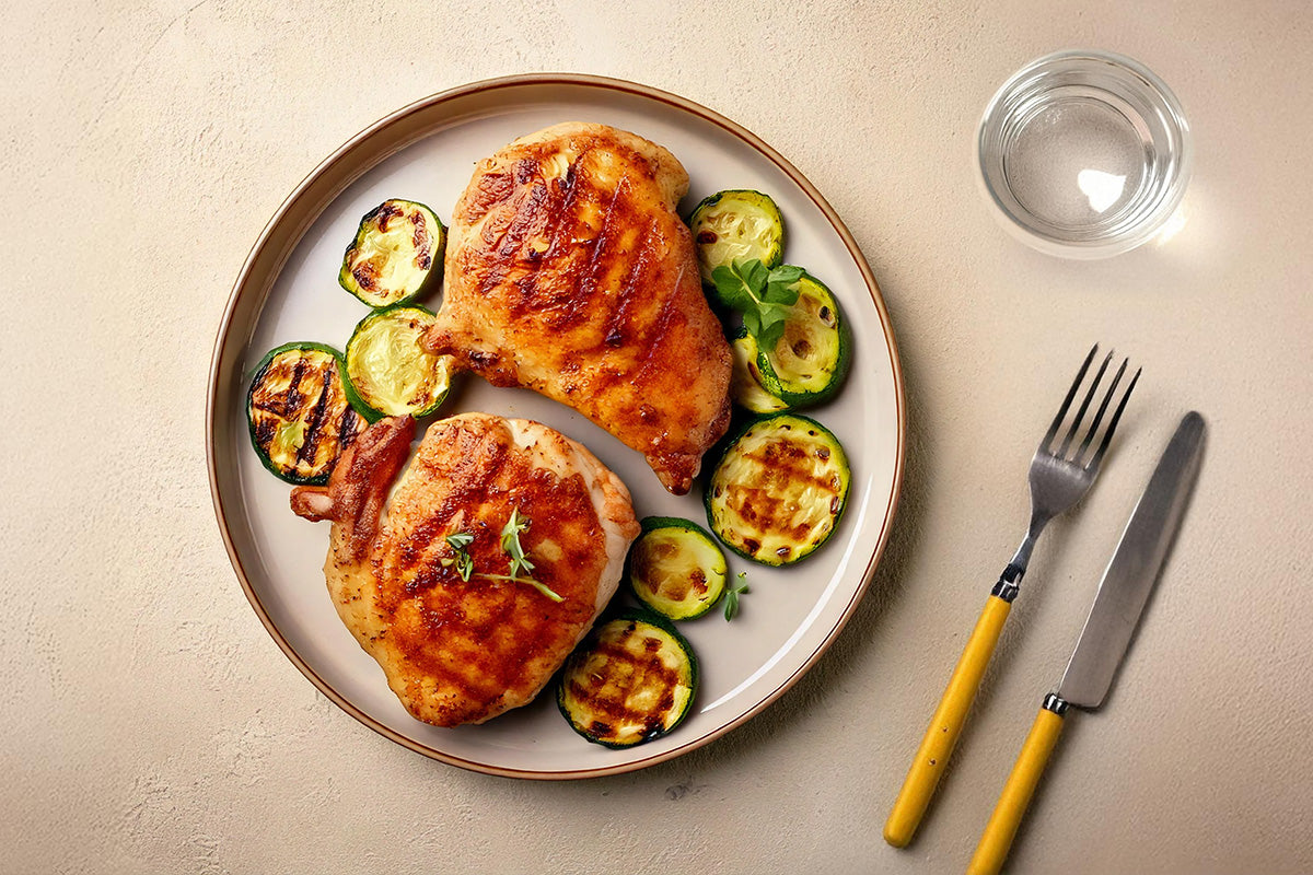 Zwei gegrillte Hähnchensteaks mit Grillgemüse aus Zucchinischeiben auf weißem Teller, daneben Besteck mit gelben Griffen und ein Glas Wasser