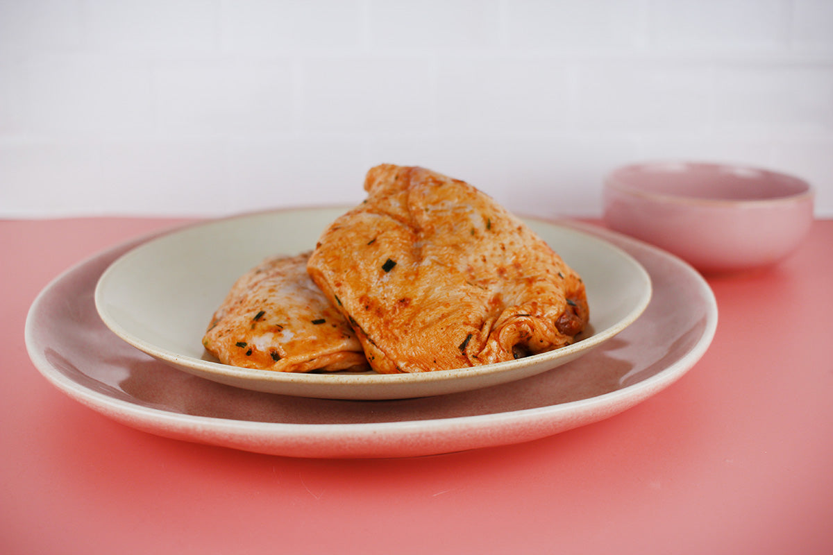 Zwei gewürzte Hähnchensteaks auf einem weißen Teller, der auf einem größeren rosa Teller steht, vor rosa Hintergrund