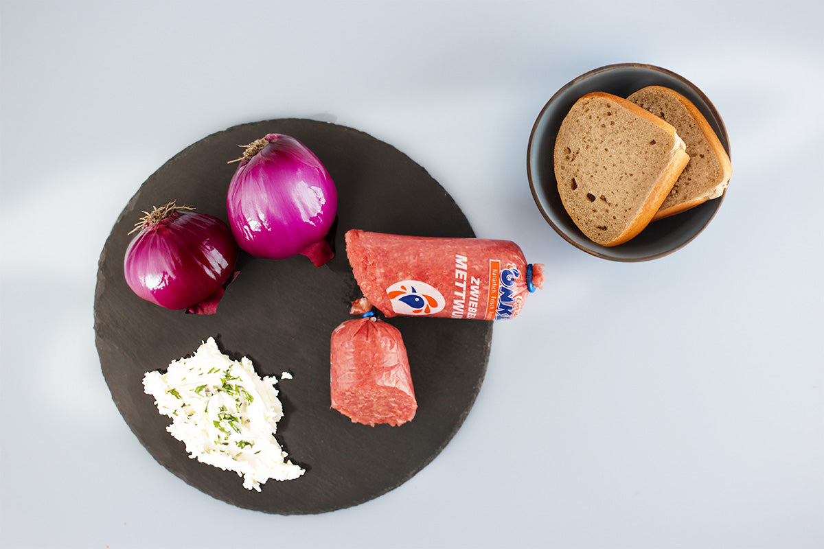 Geflügel Zwiebelmettwurst auf schwarzer Platte mit zwei roten Zwiebeln und Frischkäse, daneben zwei Scheiben Brot in grauer Schale