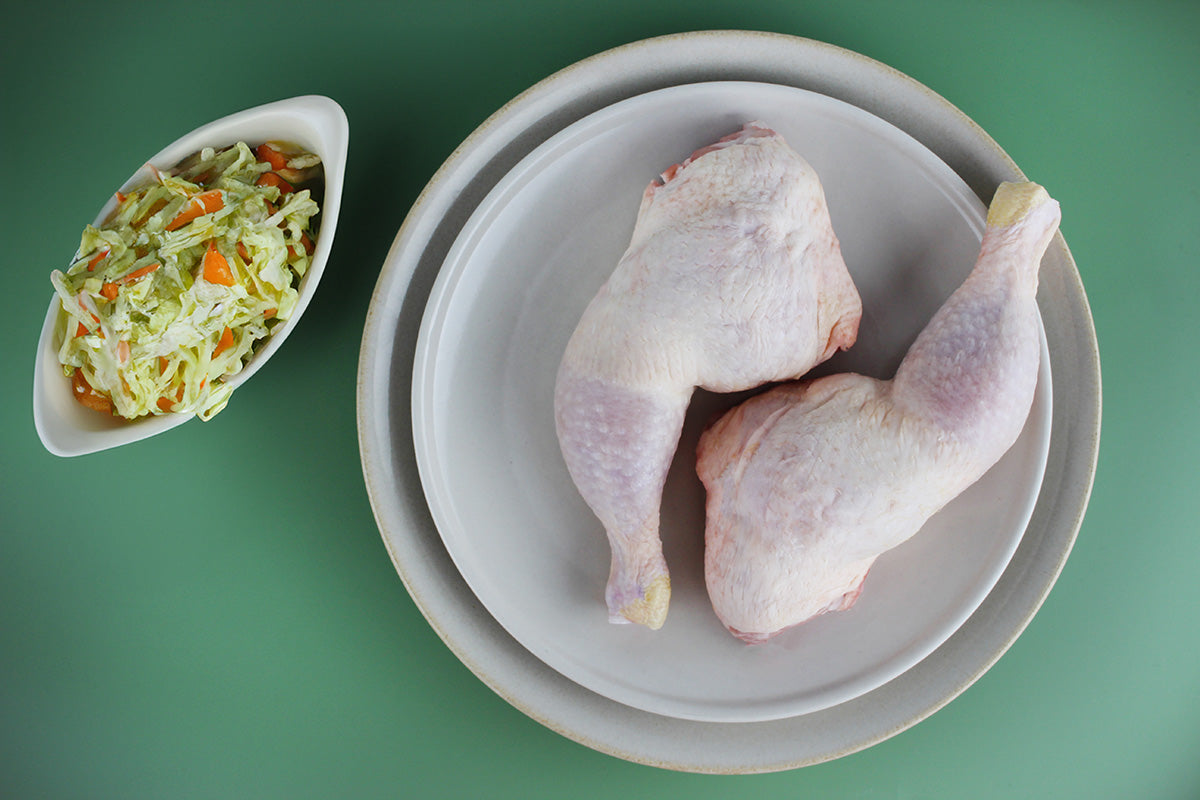 Zwei rohe Hähnchenkeulen auf einem weißen Teller, daneben eine Schale mit gemischtem Salat aus Kohl und Karotten auf grünem Hintergrund