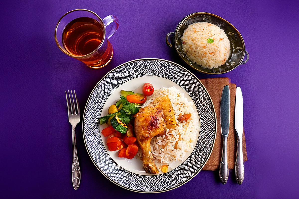 Teller mit gebratener Hähnchenkeule, Reis und gemischtem Gemüse, daneben Besteck und ein Glas Tee auf lila Untergrund, kleine Schale mit Reis