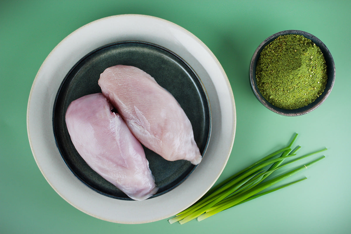 Zwei rohe Hähnchenbrustfilets auf einem schwarzen Teller, daneben grüne Kräuter und eine Schale mit grünem Gewürzpulver auf grünem Hintergrund