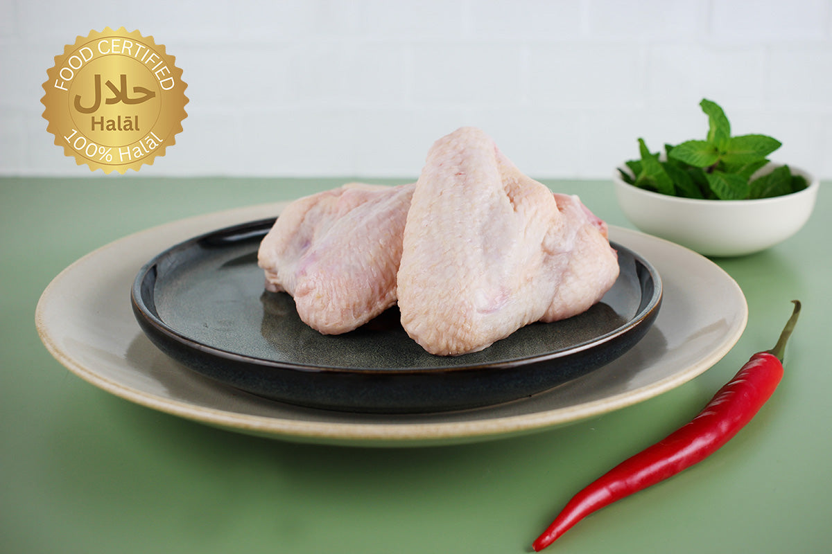 Zwei rohe Hähnchenflügel auf einem schwarzen Teller, daneben rote Chilischote und Schale mit frischer Minze, Halal-Zertifikatssiegel