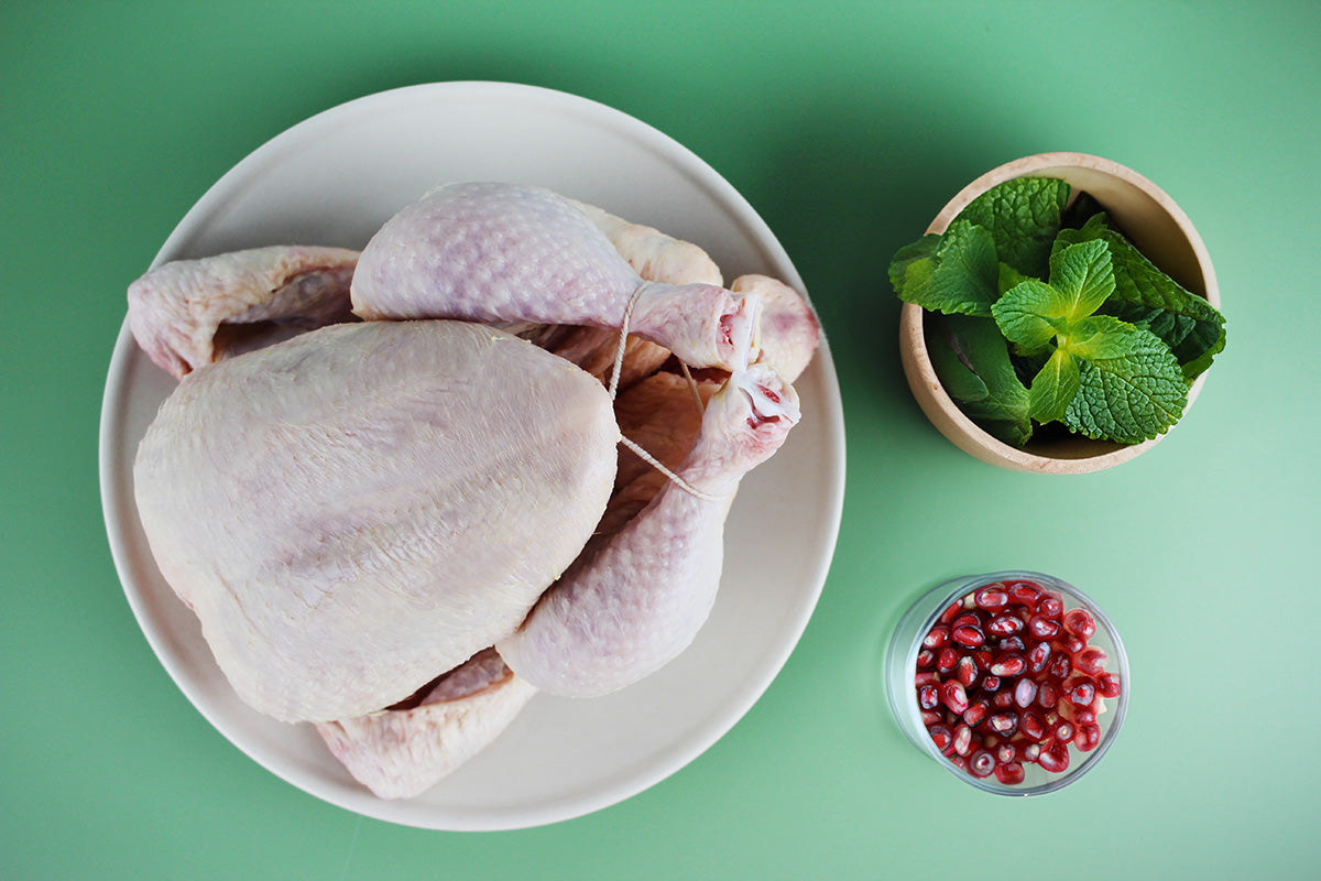 Rohes ganzes Hähnchen auf einem weißen Teller, begleitet von einer Schale mit frischen Minzblättern und einer Schale mit Granatapfelkernen, auf grünem Hintergrund.