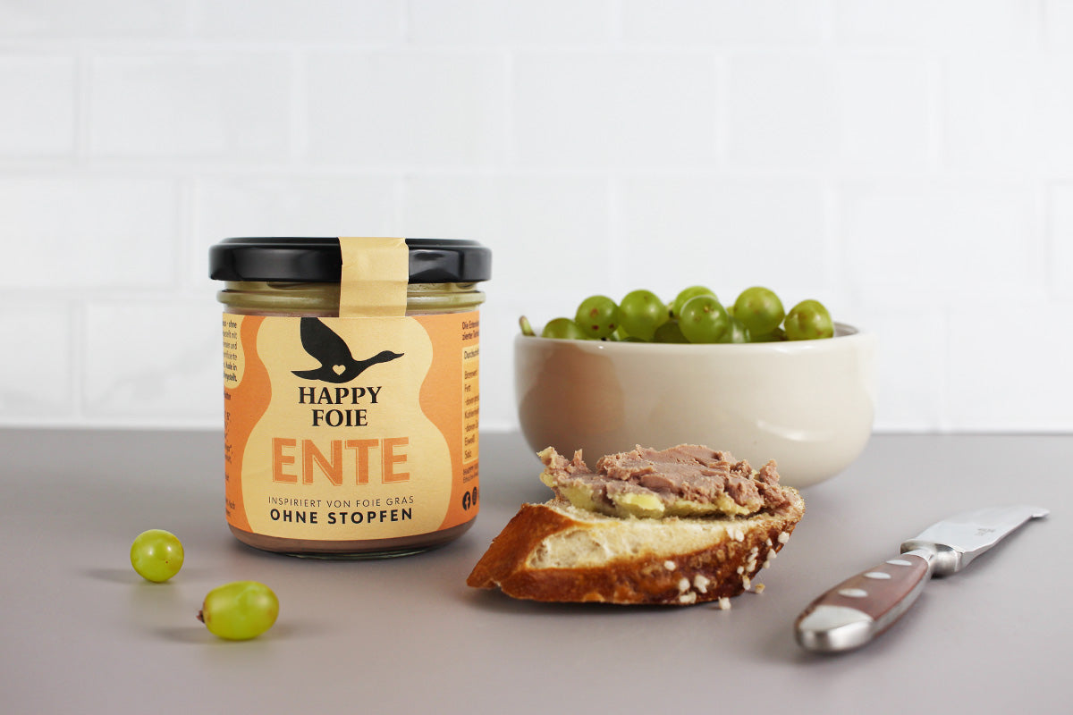 Glas Happy Foie Ente Entenpastete, daneben ein Stück Brot mit Pastete, eine Schale mit grünen Trauben und ein Messer auf grauem Untergrund