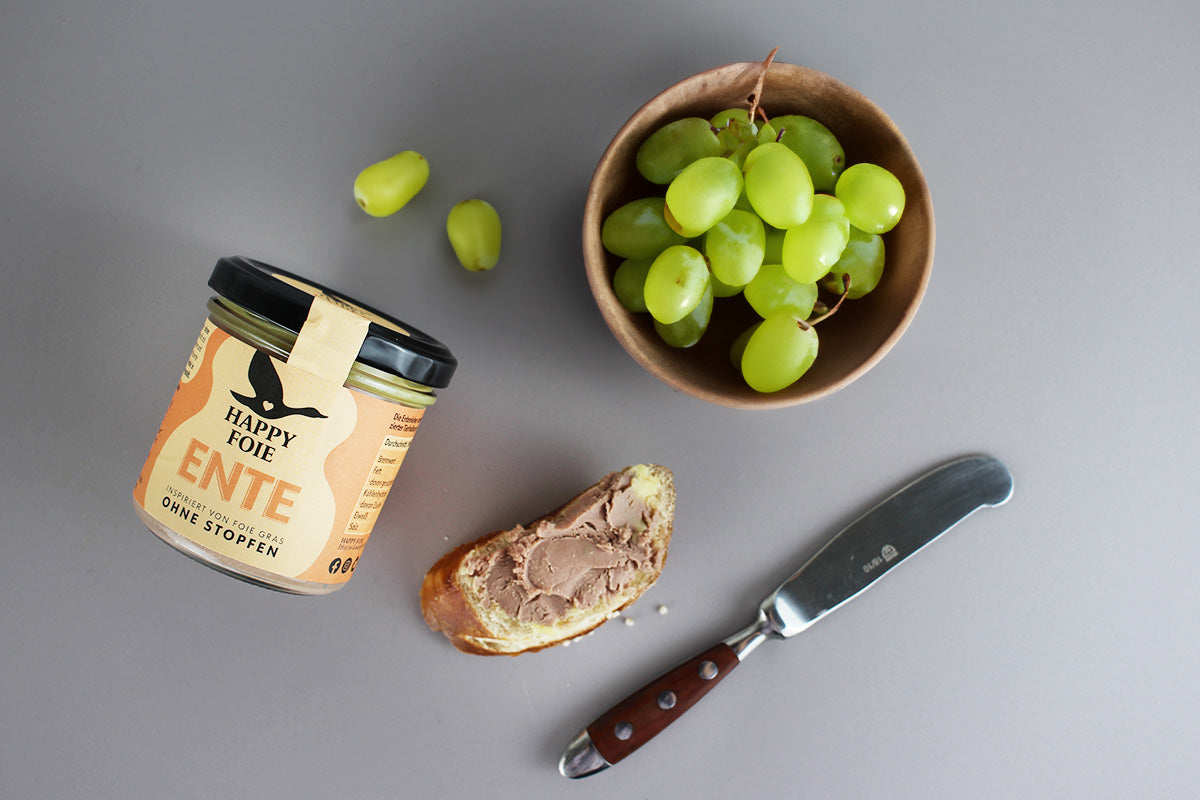 Glas Happy Foie Ente Pastete, Scheibe Brot mit Pastete, Messer mit Holzgriff und Schale mit grünen Trauben auf grauem Untergrund