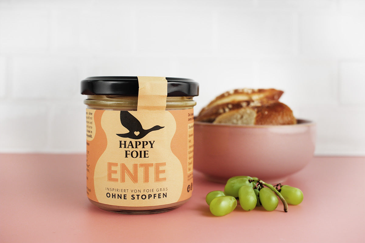 Glas mit Happy Foie Entenpastete, daneben grüne Trauben und eine Schale mit Brot