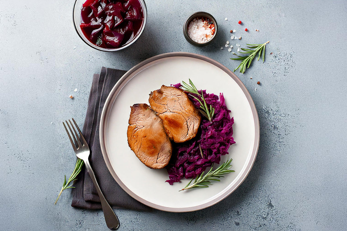Zwei Scheiben gebratene Hirschnuss auf einem Teller mit Rotkohl und Rosmarinzweigen, daneben eine Schale mit Rotkohl und eine kleine Schale mit Salz und Pfeffer