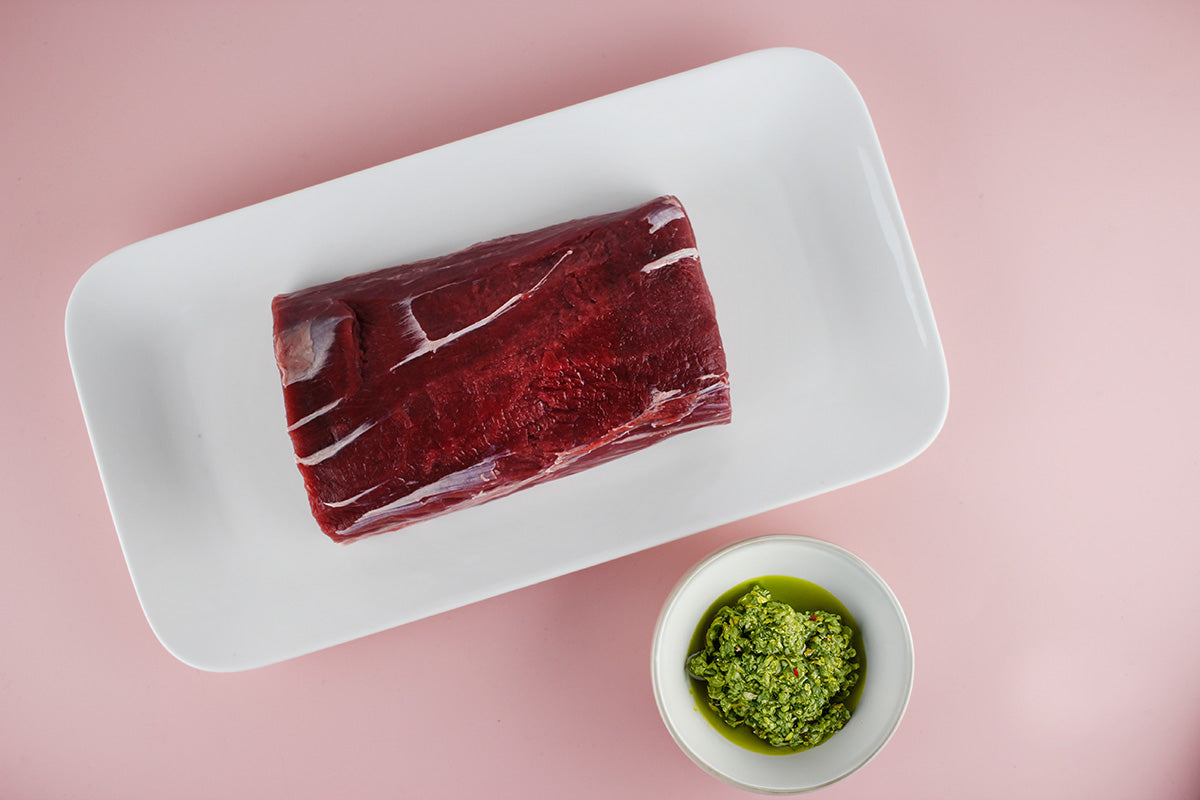 Roher Hirschrücken auf weißem Teller neben einer kleinen Schale mit grüner Kräuterpaste auf rosa Untergrund