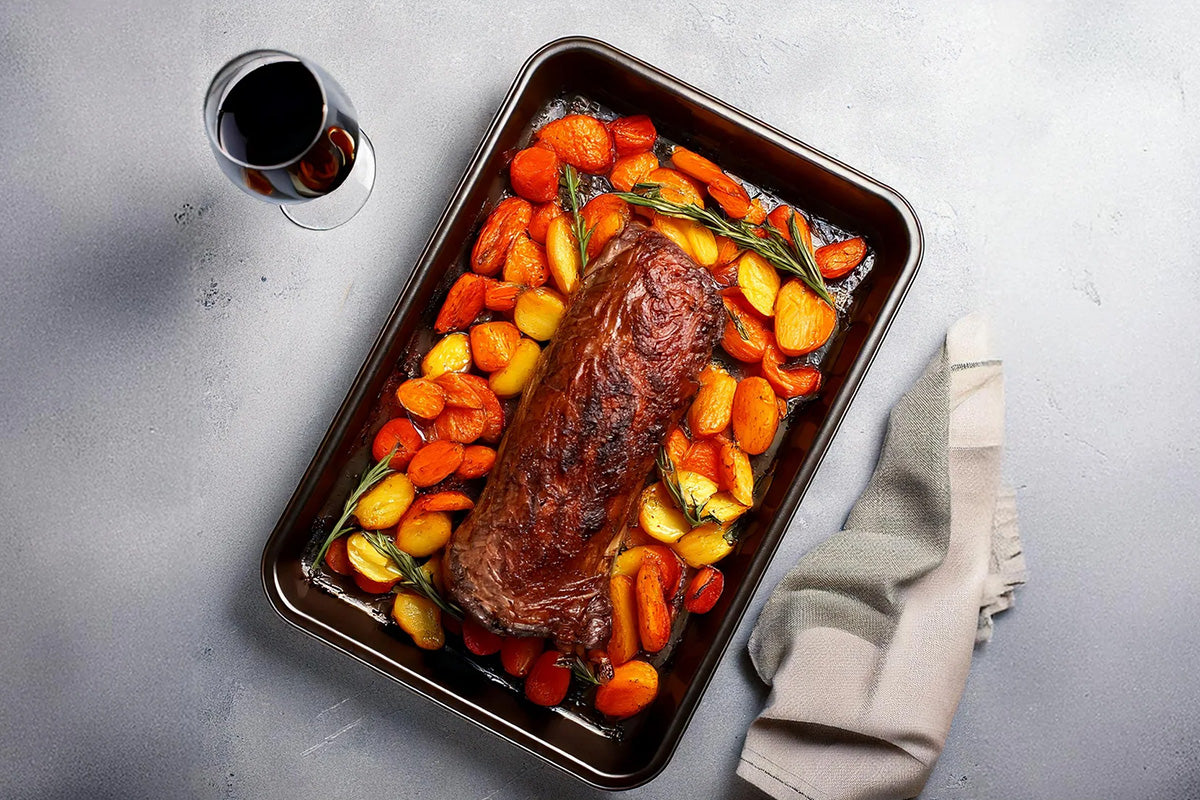 Gebackener Hirschrücken mit gerösteten Karotten und Kartoffeln in einer Bratform, daneben ein Glas Rotwein und ein graues Küchentuch