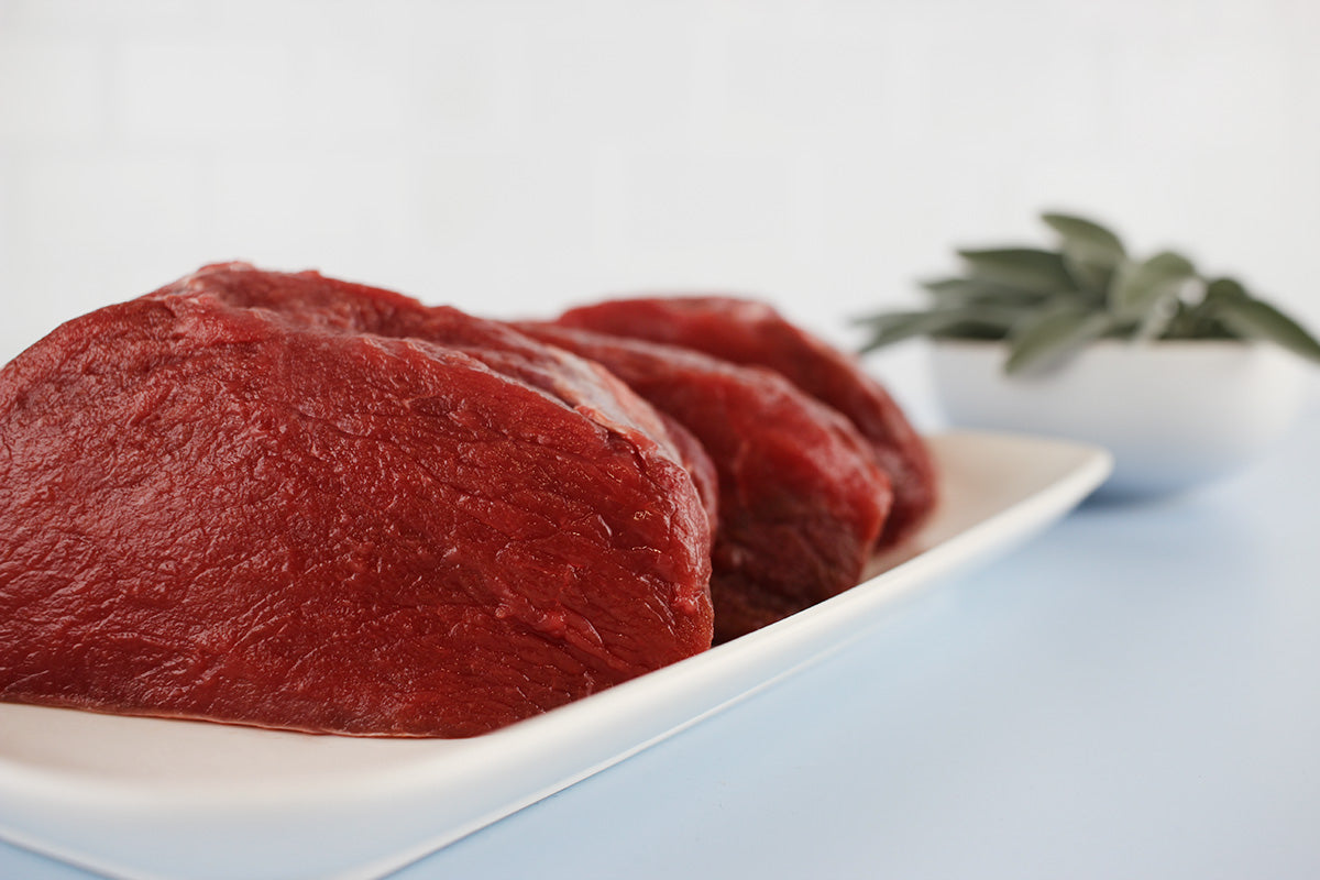 Drei rohe Hirschsteaks auf weißem Teller, unscharf im Hintergrund Schale mit frischen Kräutern