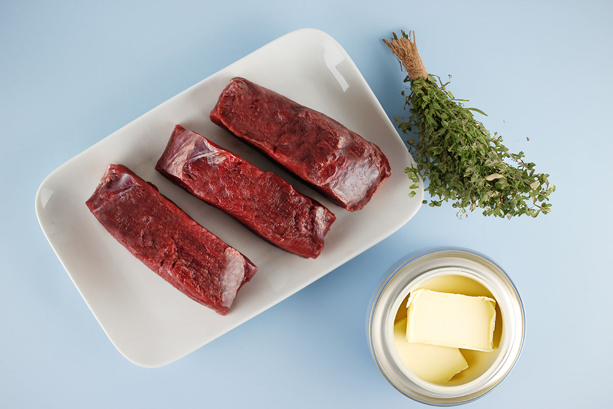 Drei rohe Hirschsteaks auf weißem Teller, daneben Butter in Glas und ein Bund getrockneter Kräuter auf blauem Untergrund