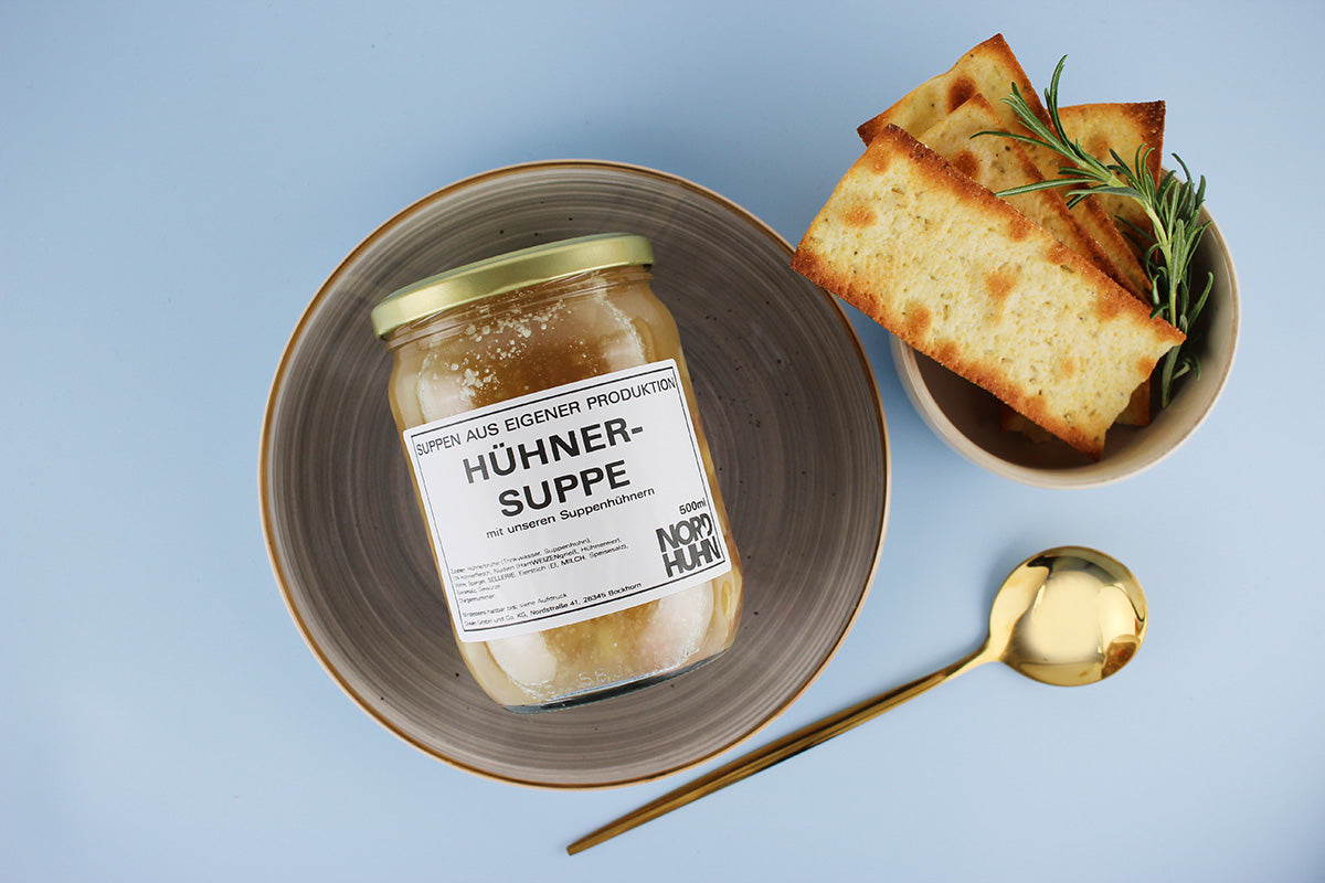 Ein Glas Hühnersuppe mit Aufschrift 'Hühnersuppe' auf einem grauen Teller, daneben ein Becher mit geröstetem Brot und Rosmarinzweig, goldener Löffel auf blauem Hintergrund