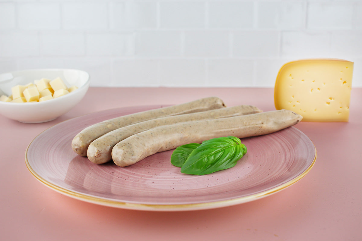 Drei helle Käse-Geflügelbratwürste auf rosa Teller mit Basilikumblatt, daneben Stück Käse und Schale mit Käsewürfeln