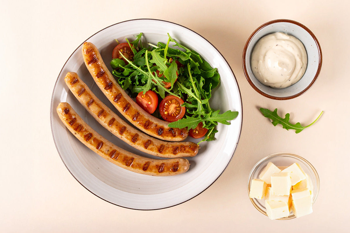 Drei gegrillte Geflügelwürstchen auf Teller mit Rucola und Kirschtomaten, daneben Schüssel mit weißer Sauce und Schale mit Käsewürfeln