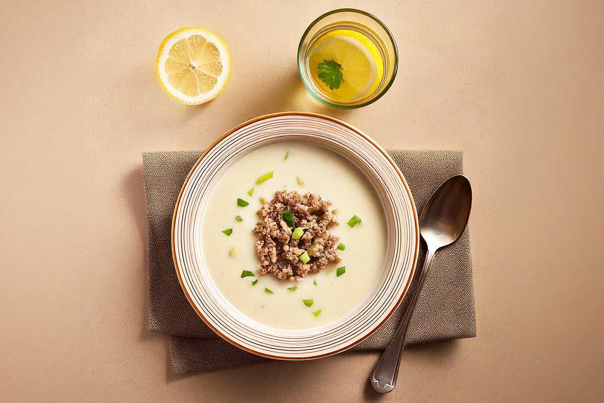 Schale mit cremiger Käsesuppe, garniert mit Hackfleisch und Frühlingszwiebeln, daneben halbe Zitrone und Glas Wasser mit Zitronenscheibe und Petersilie